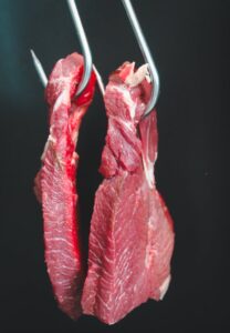 Two raw beef cuts hanging on metal hooks against a dark background.